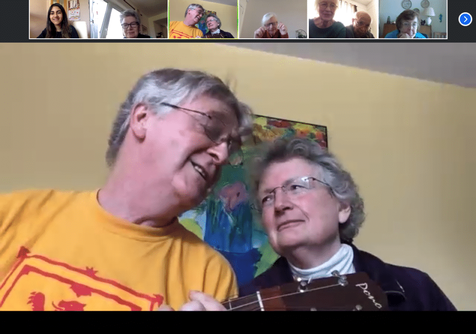 A screenshot of a video call, a large box with two older people with grey hair sat together, one playing a guitar, with a row of smaller boxes at the top showing the other callers
