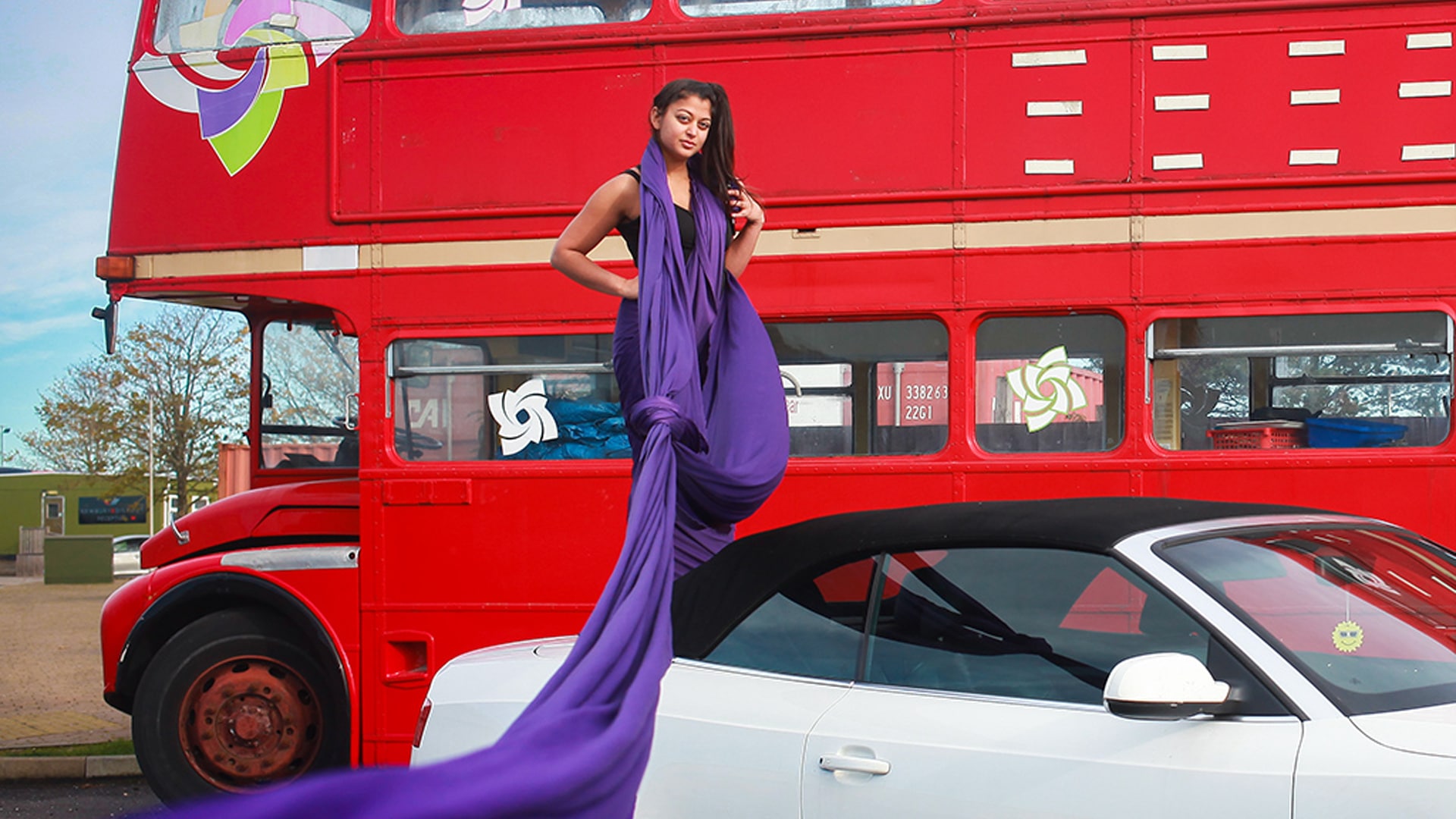 A person wearing a purple dress with a lush long purple train from their neck stood on the back of a white soft top convertible car in front of a classic red double decker bus