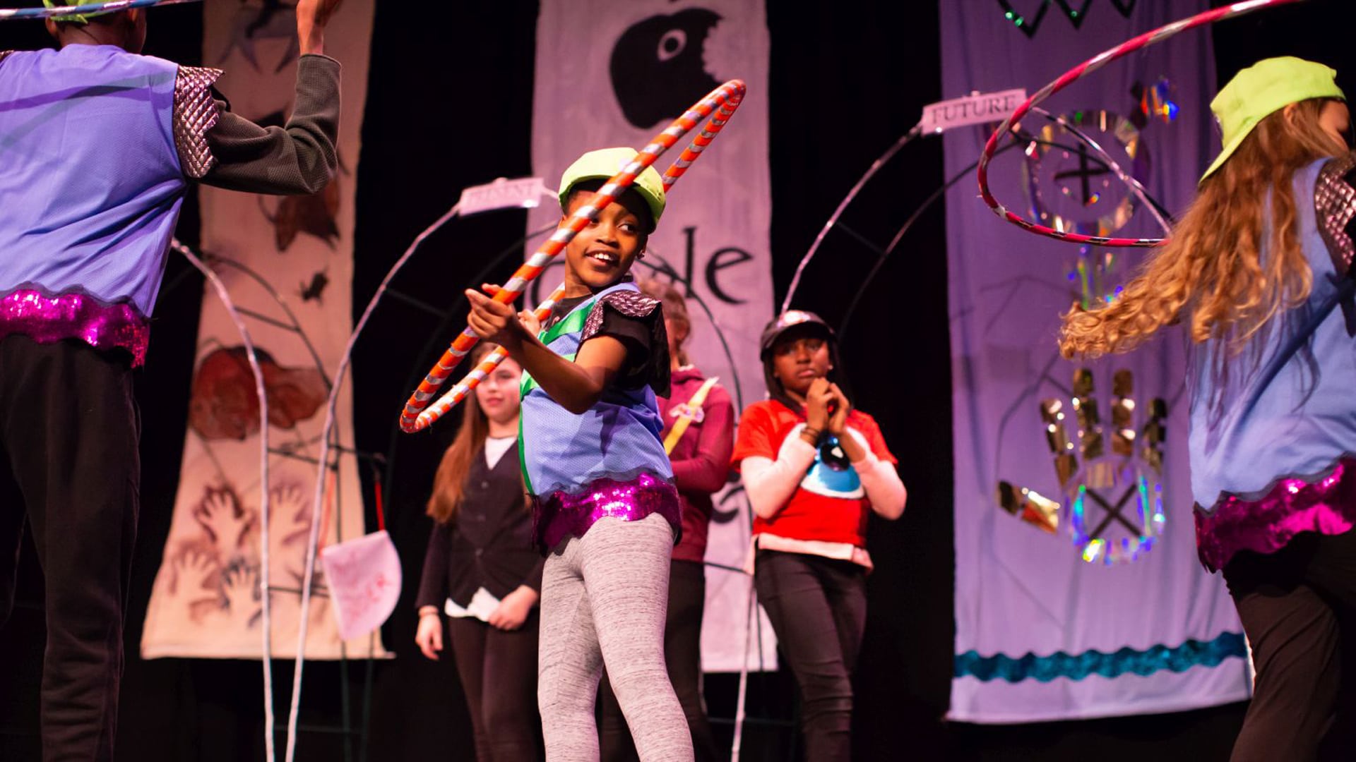 Young performers on stage, one in the centre with a hula hoop