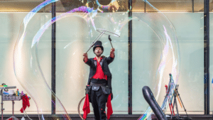 The Bubble Wizard performing on-stage, both arms raised as he makes a massive bubble in the shape of a heart
