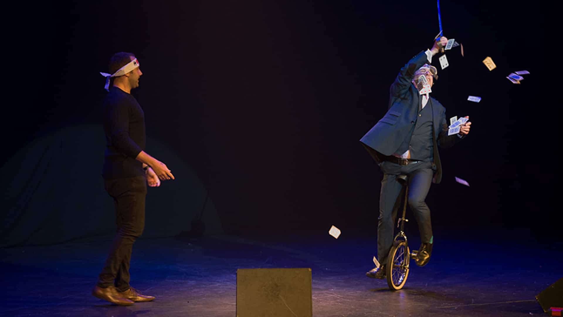 Christian Lee performing on stage, riding a unicycle, and dropping playing cards all around him. An audience member stands on stage next to him looking on