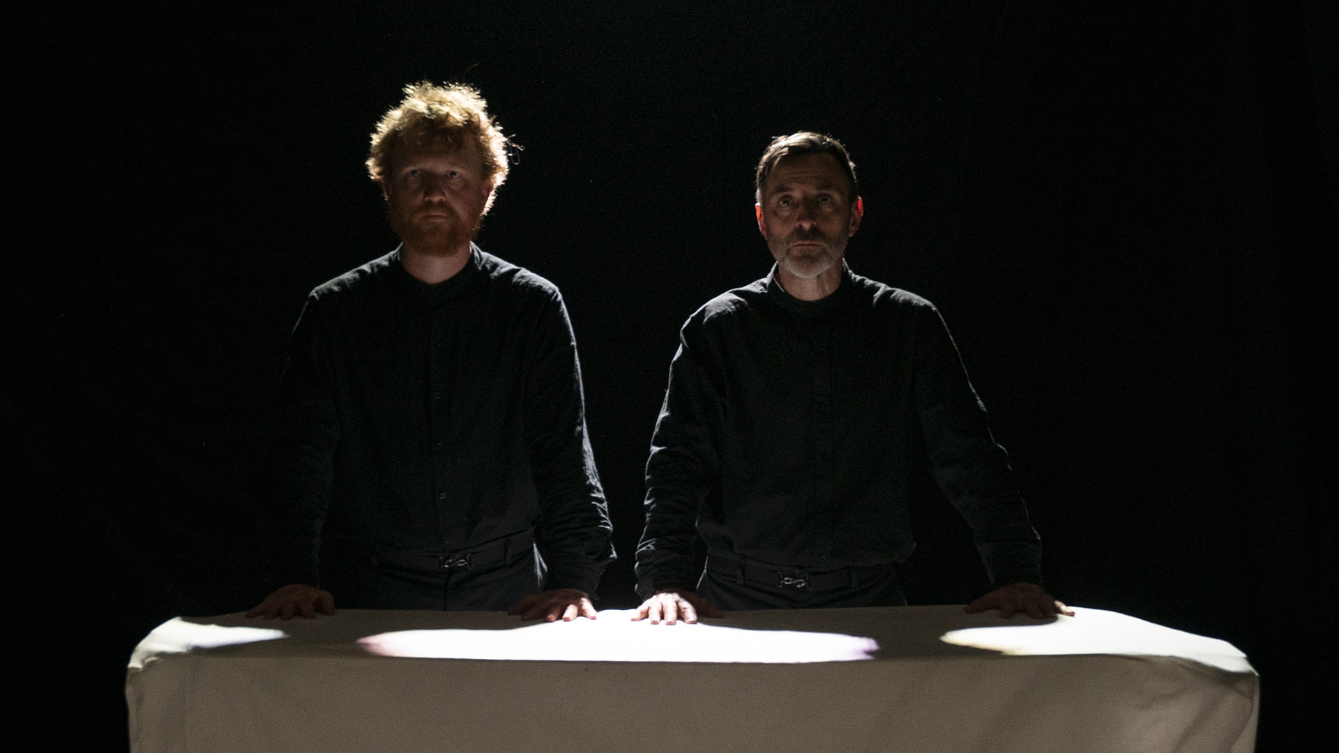 Dik Downey and Adam Blake stand behind a table, covered in a white tablecloth. They are cast in shadows, their hands pressed onto the tabletop