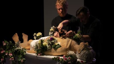 A body lies prone on a table covered in funeral flowers. Dik Downey and Adam Blake stand over it.