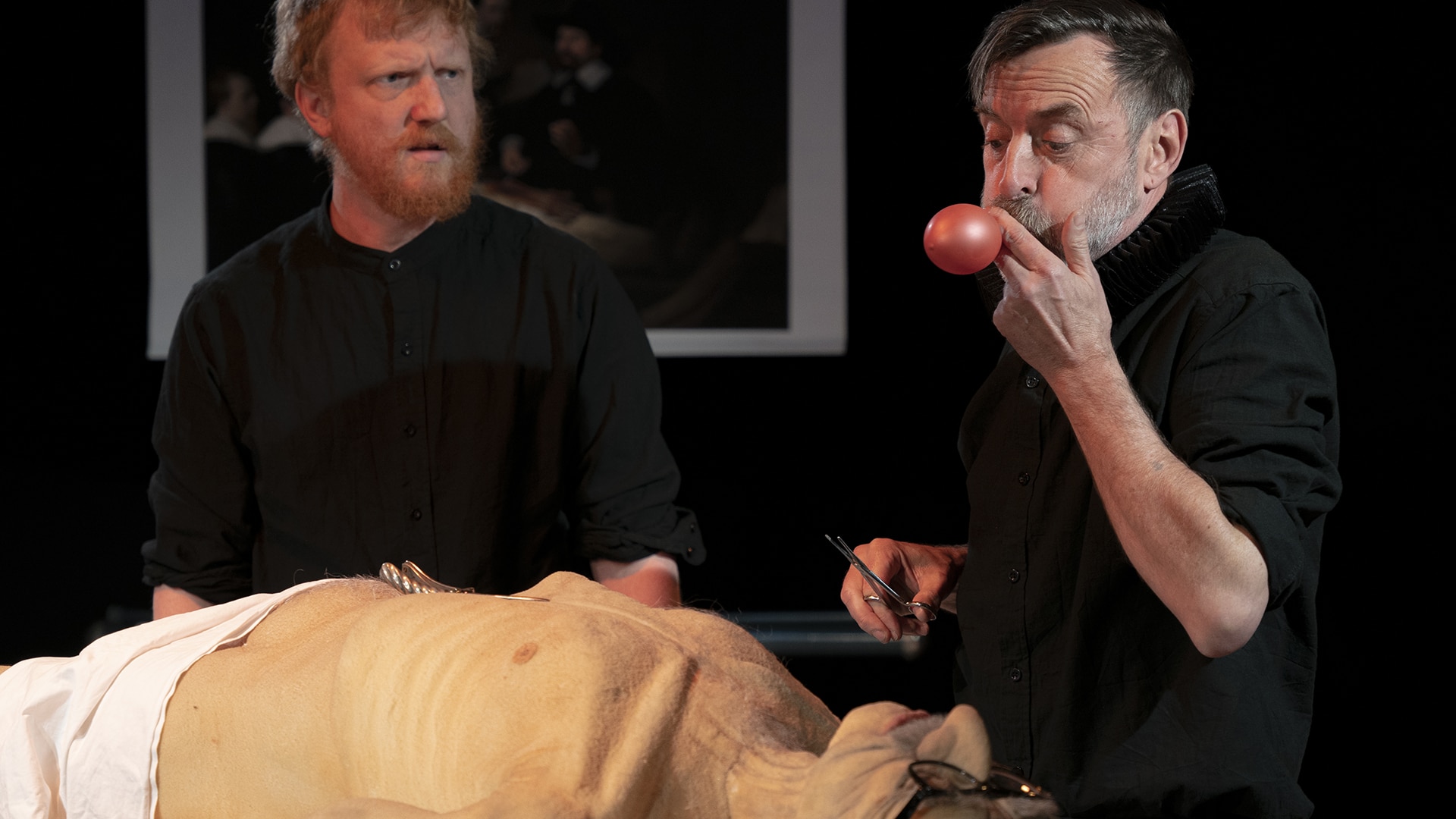 A body lies prone on a table. Dik Downey and Adam Blake stand over it. Adam watches Dik as he blows up a balloon while holding a pair of surgical scissors