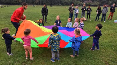 A group of children stand in a circle outside holding onto a multicoloured parachuts. A man in a red top holds one side, guiding their activity