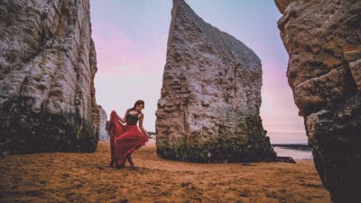 A dancer moves on a deserted beach, surrounded by towering rocks. She wears a long red skirt which swirls around her legs