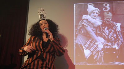 Dr Marisa Carnesky speaks into a microphone while standing beside the projected black and white photo of two clowns