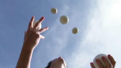 Viewed from below, hands juggle three balls against the blue sky