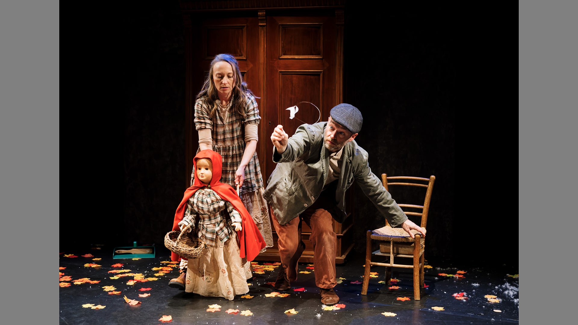 A photo of two actors on stage. A woman moves a puppet of a girl wearing a red cloak and carrying a basket. A man leans foward, balancing one hand on a wooden chair. He is wearing a green jacket and grey flatcap