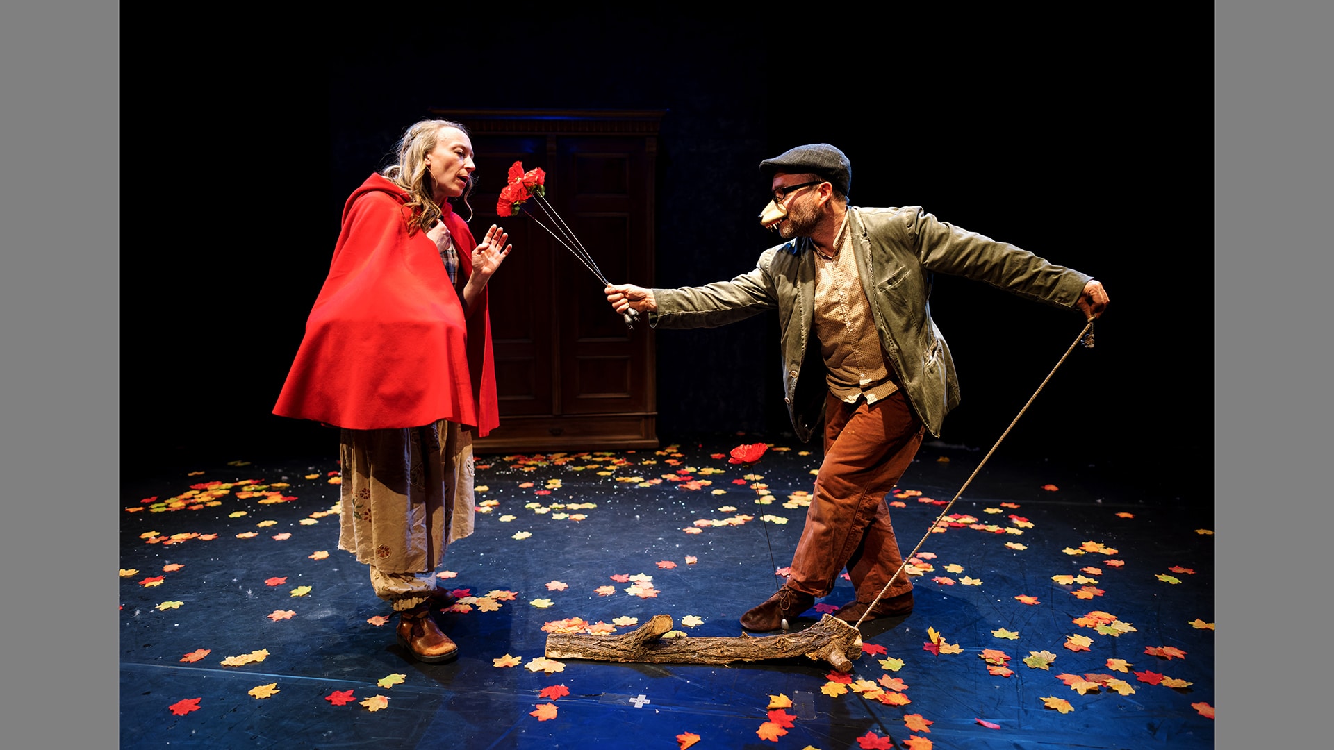 A man wearing a green jacket and flatcap offers a bouquet of red flowers to a woman wearing a red cloak