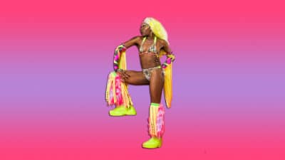 Symone poses with one leg raised. She wear yellow trainers, a yellow bikini and has a yellow wig. She has pink leg warmers and arm warmers, with yellow and pink fringes on them. She looks strong and powerful