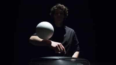 A man hidden in shadows balances a white ball on his forearm