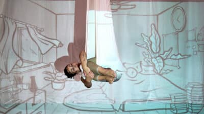 Performer curls up, like a sleeping baby, in a hanging sling. An image is being projected over her. It shows a bedroom, the open window, the curtains blowing in the wind