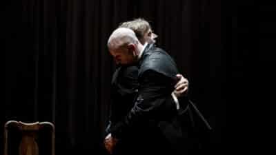 Two older men dance together, wrapped in each other's arms, baring each other's weight