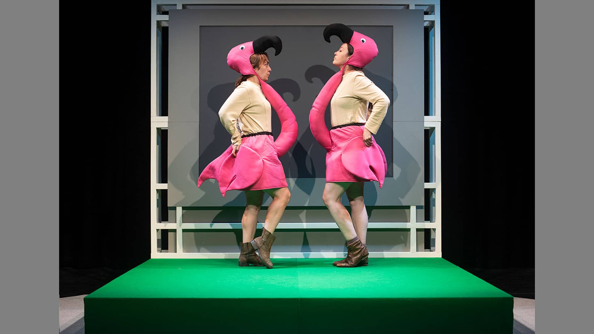 The two performers are dressed as flamingos. They face each other on stage, their arms bent like wings