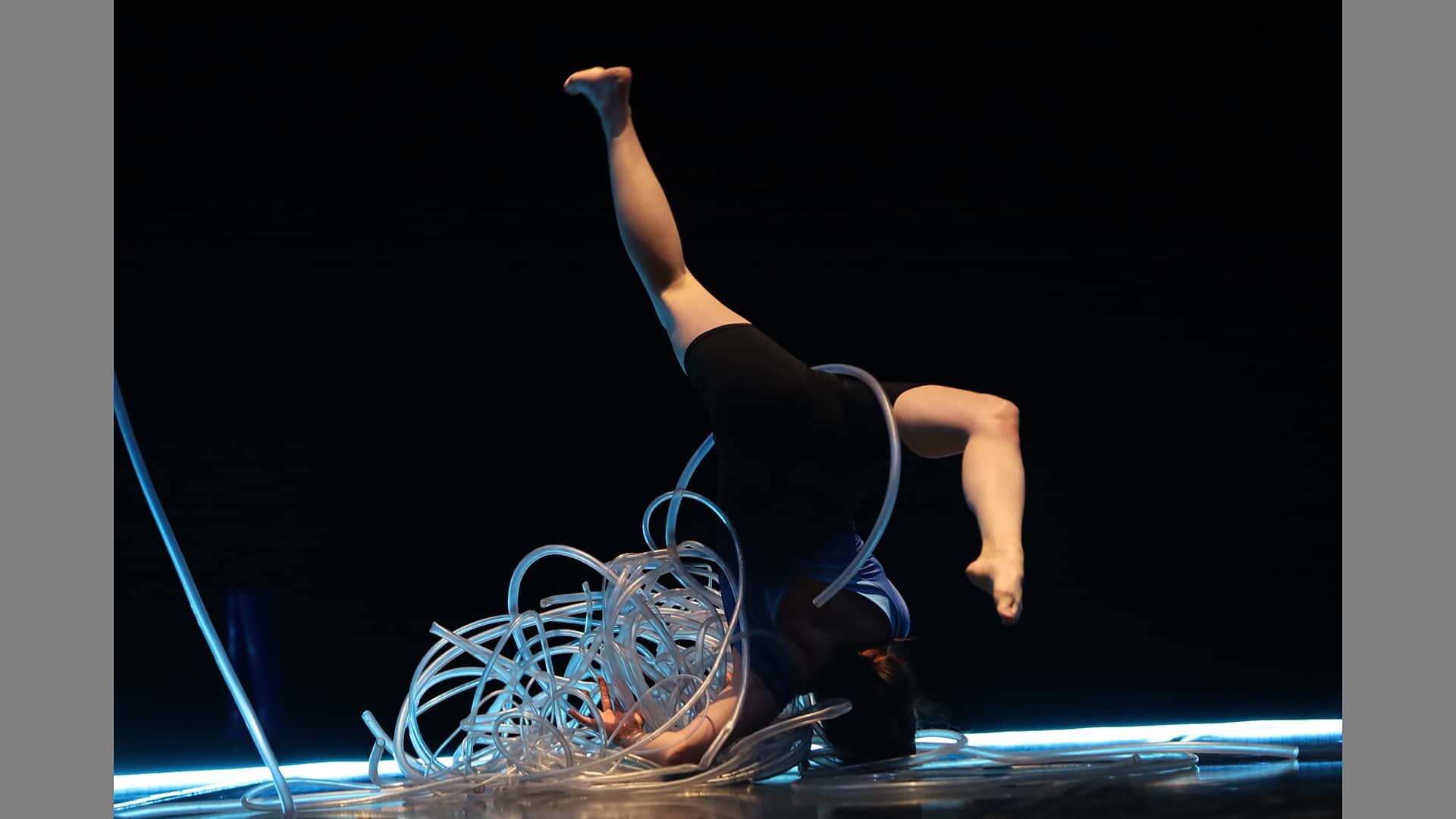 Performer is upside down, her legs in the air, bent. Her face is hidden behind a mound of plastic tubing