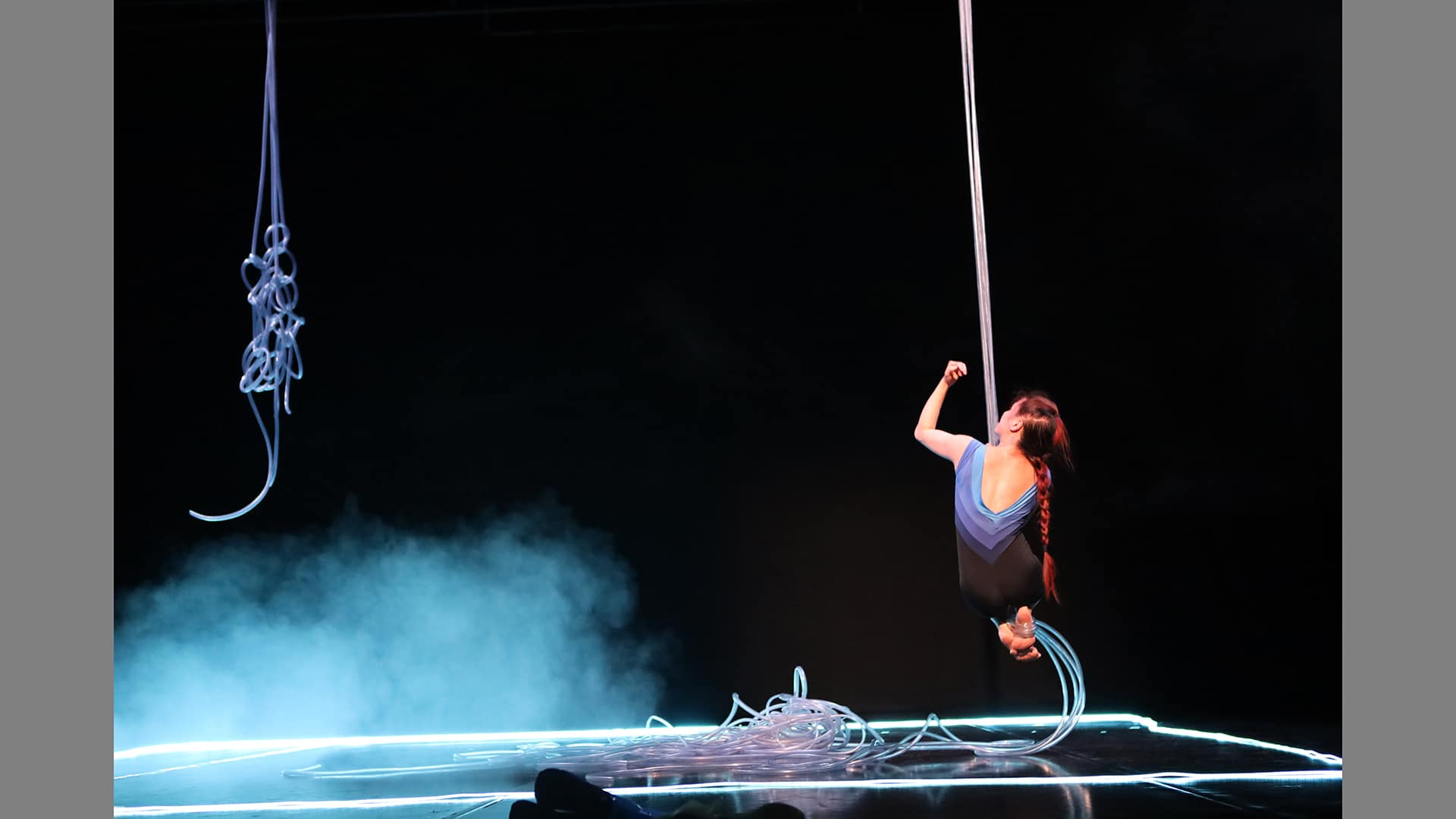 A performer swings around the stage, her back to the camera, her feet tucked up under her, using plastic tubing as a rope. Her long red hair is tied back in a plait