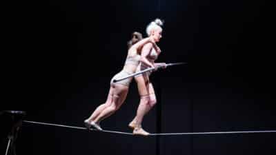 Two performers cross the tightwire. One holds a balance bar. The other clings to her from behind