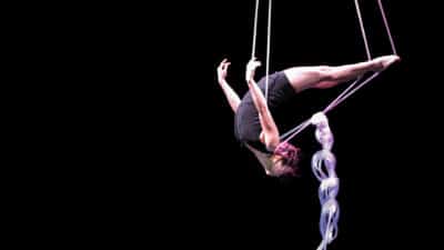Performer swings above the stage using plastic tubing as rope - she's upside down, her legs stretched out behind her, holding out the tubing from her body. Her back is arched in an upside down swan dive