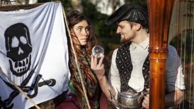 A pirate holds a crystal ball in one hand and a silver teapot in the other. Another performer is half hidden by a pirate flat.