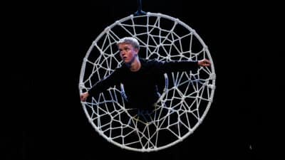 A performer emerges from a huge circle covered in a net which looks like a spider's web. Half their body is through. Their arms grip the sides of the circle