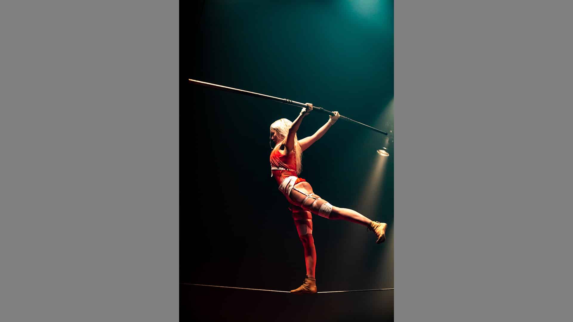 A photo of Hanna Moisala on the tightwire. Her back leg is lifted in an arabesque. Her arms are above her head, holding the balance bar above her