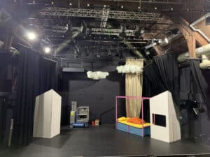Photo showing the set of Jack and the Beanstalk while it's being built. There is a four poster off to the left, with pink posts and yellow sheets. There are two walls either side. Both are white, waiting to be painted. A couple of fluffy clouds hang from the ceiling. The lighting rig is visible above the clouds