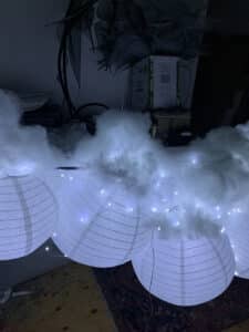 A photo of white paper lanterns stuck together in a cloud shape. They have fairy lights inside, and they are half covered with a fluffy material which looks like white cotton wool