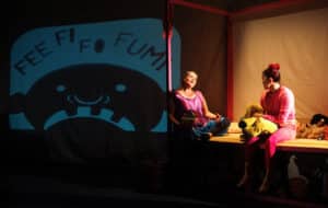 Performers playing mum and Jackie sit on Jackie's bed, as mum tells a bedtime story. Behind them is a shadow puppet of a giant with scary teeth showing. Above the giant in big letters it says Fee Fi Fo Fum. Jackie holds a yellow toy dog on her lap