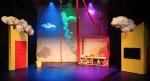 The Jack and the Beanstalk set. There is a red four poster bed near the middle, with red aerial silks tied to the posts. There are yellow walls on either side of the stage - one with a red door, the other with a window. On the stage is a yellow bucket, out of which grows a rope beanstalk. Fluffy clouds hang above the set