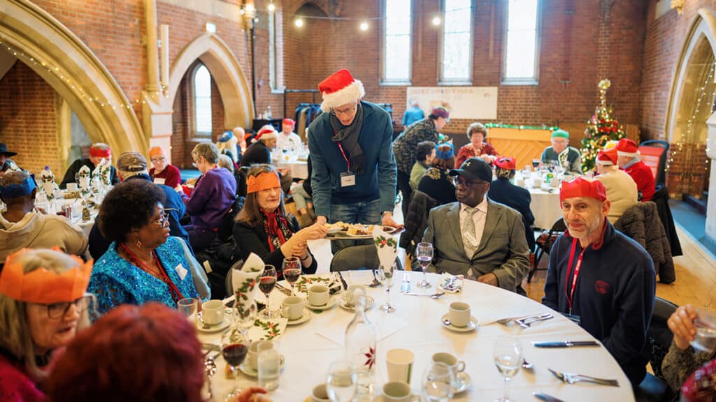 A table full of people being served dinner on Christmas Day. A volunteer is serving a tray full o f starters