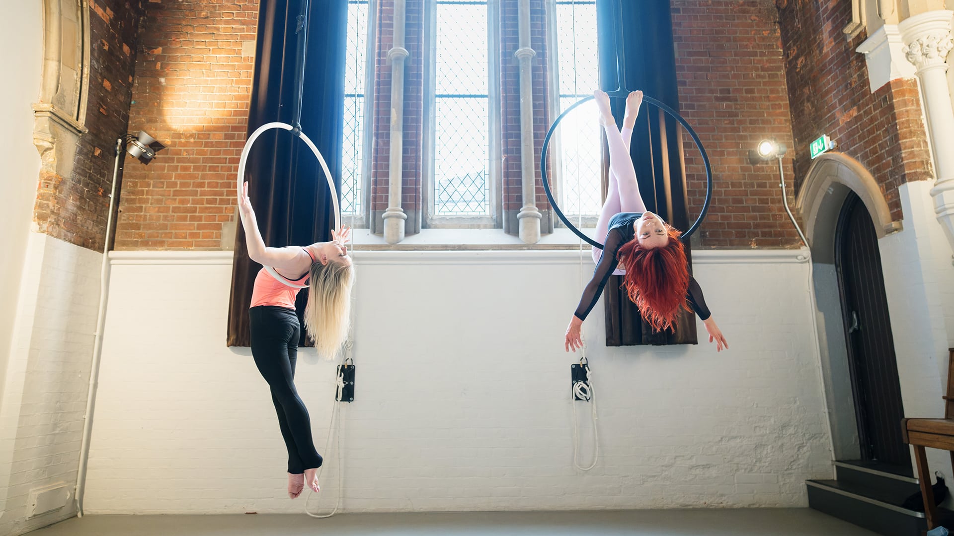 Two aerial artists hand from hoops in the rehearsal studio. One lies on her back in the hoop, her legs crossed above her, her long red hair hanging down. The other is in a back bend, her blonde hair flying out behind her
