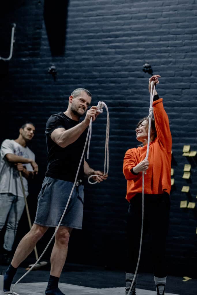 A photograph of three performers on stage. The two in the foreground each hold lengths of rope in their hands The one in the background leans on a broom and watches