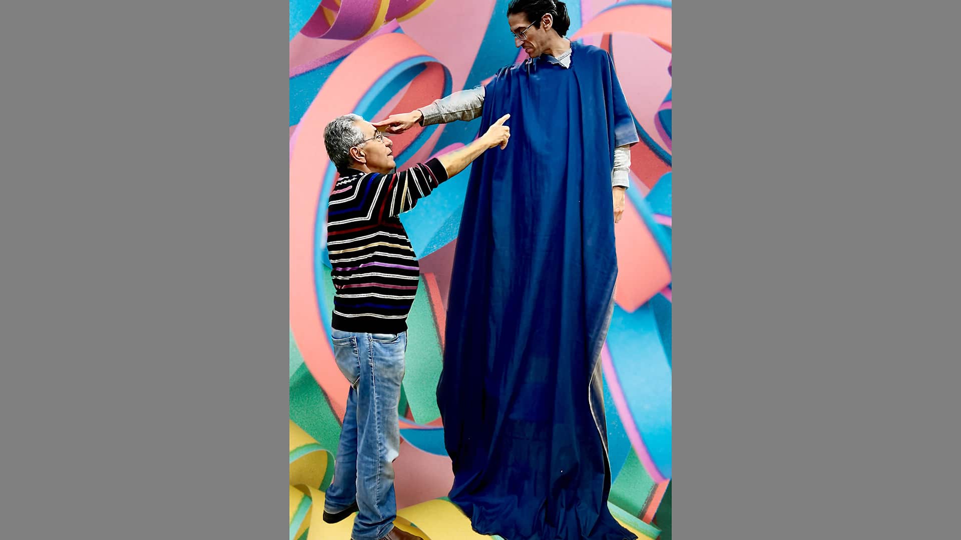 Two figures point at each other. One a man wearing a stripy jumper and jeans, the other much taller, draped in dark blue cloth