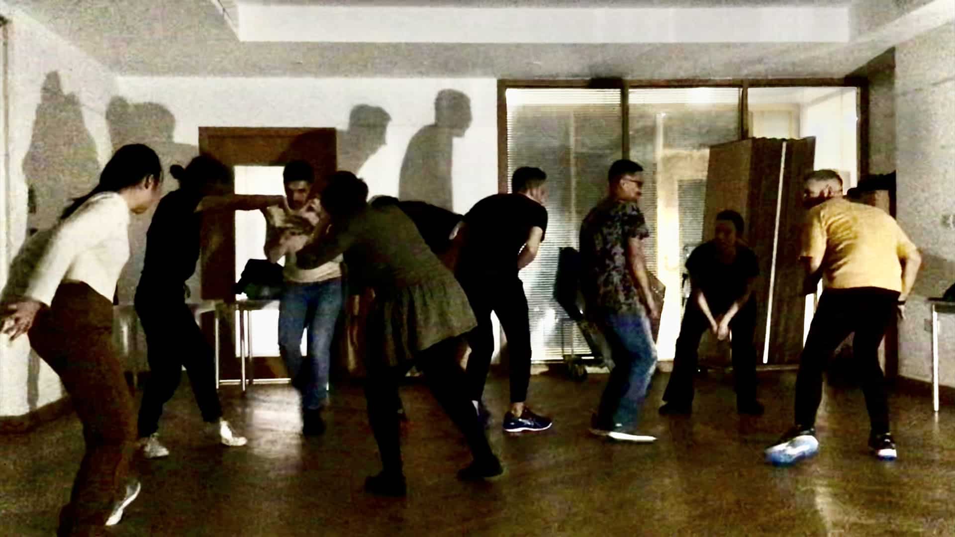 Eight performers move around a small rehearsal room. They seem to be caught in the act of sneaking about