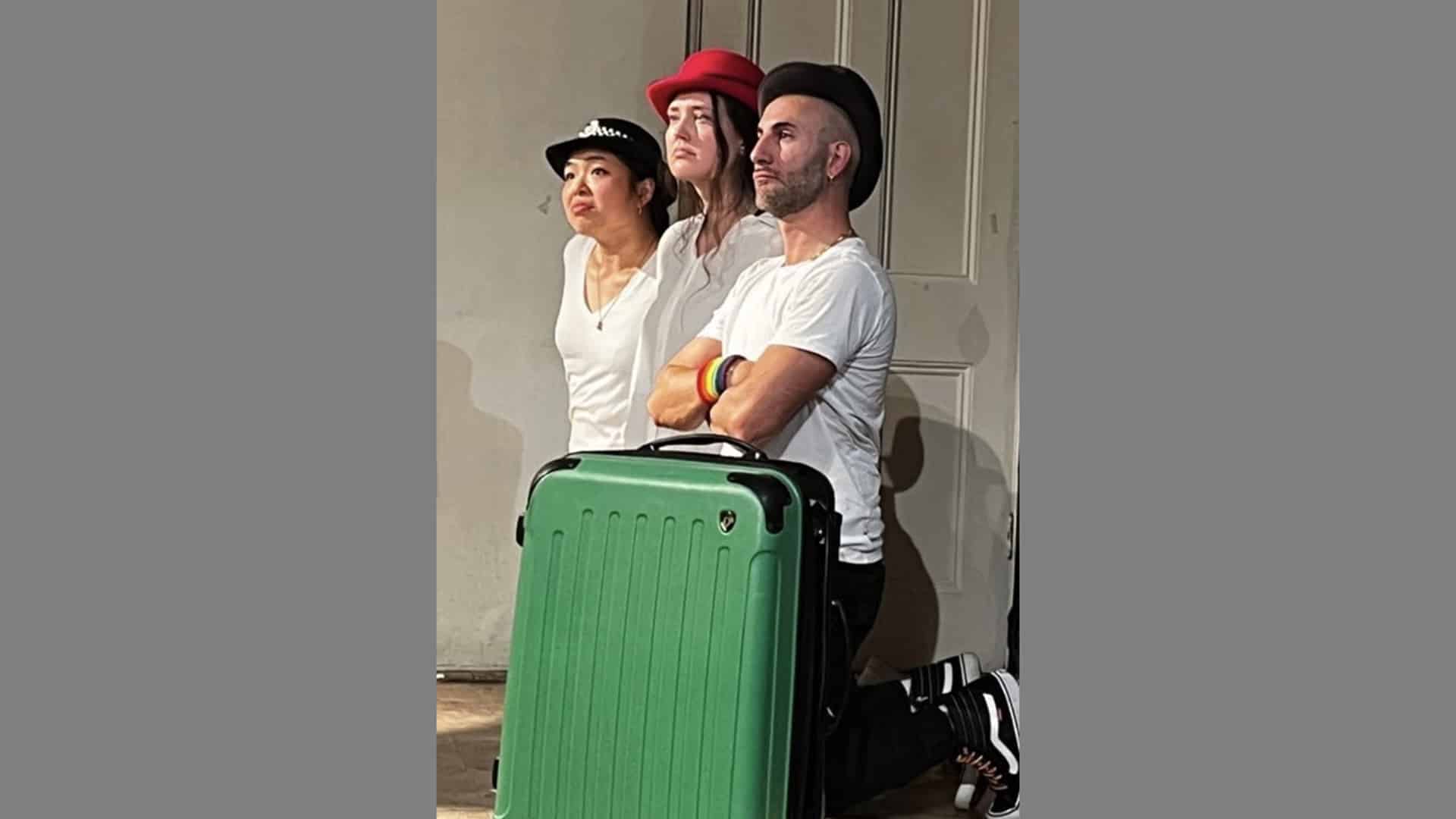 Rehearsal photo of three performers standing close to each other behind a green suitcase. They each wear different hats: red bowler, black bowler and police hat.