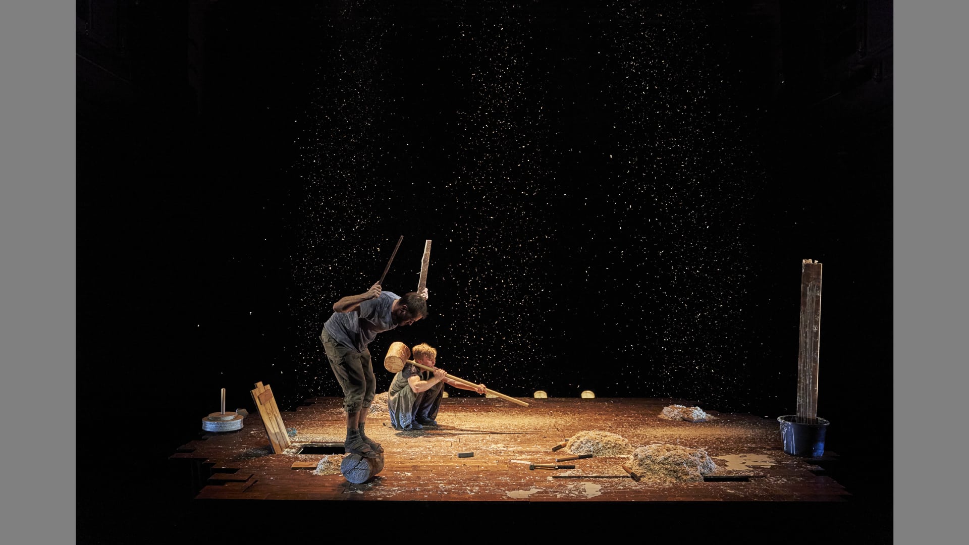 A performer balances in a rolling log, while another crouches down holding a massive hammer