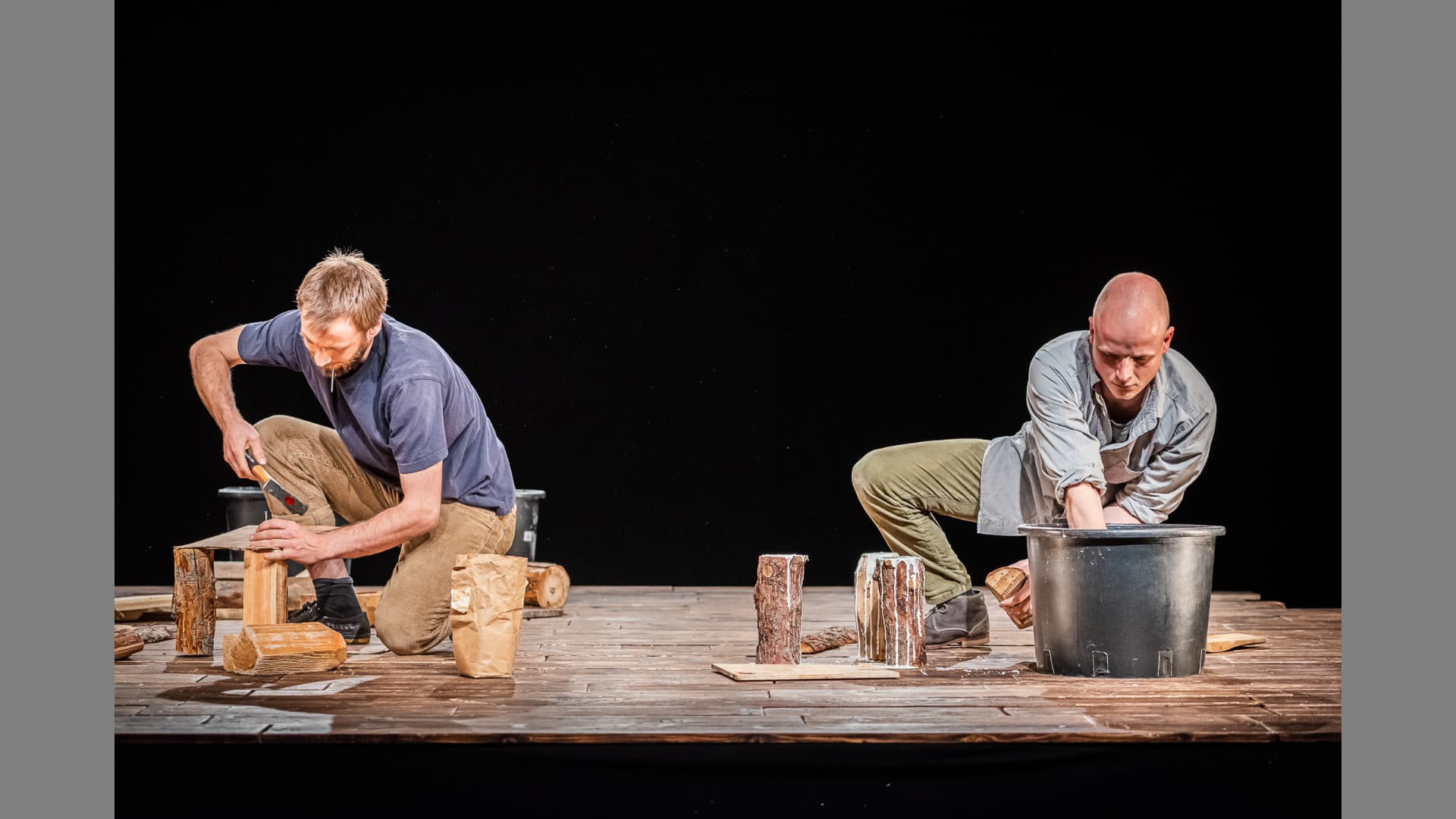 A performer crouches on the stage while doing woodwork. Another performer reaches deep into a metal bucket