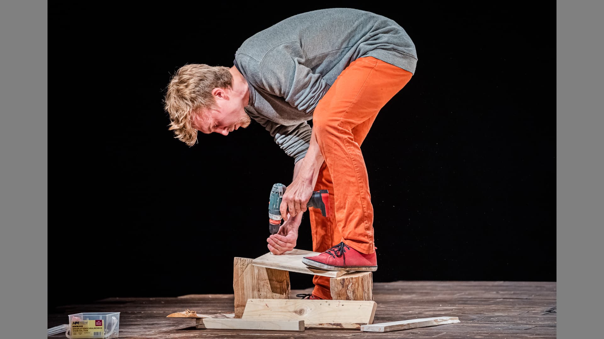 A performer leans down to screw together pieces of wood