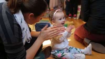 A baby sits on the floor, clapping along to music, while their mum claps with them