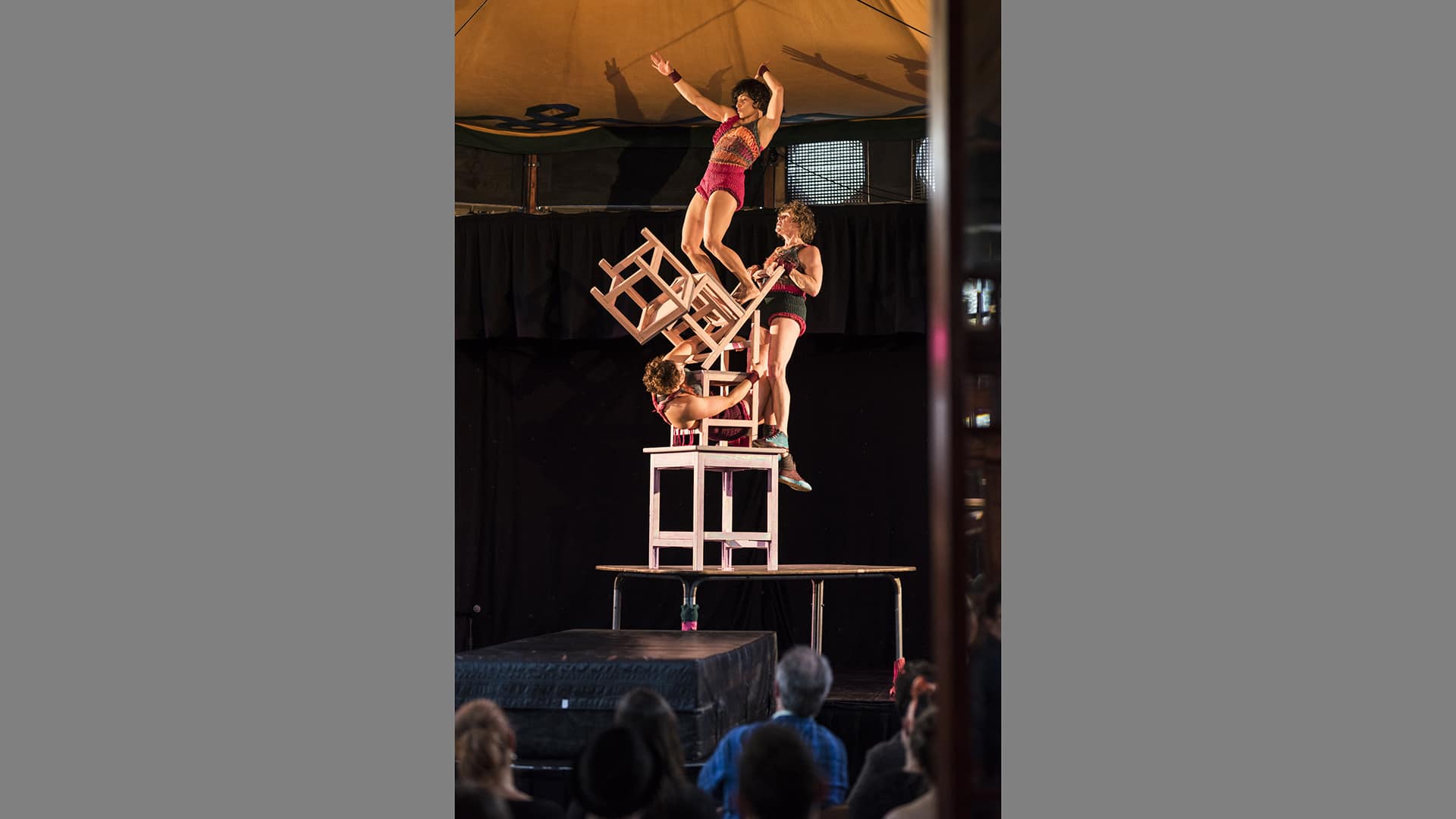 A woman balances on a tower of chairs, being held up by two other women