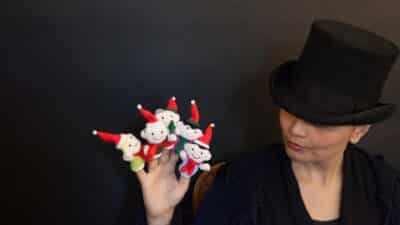 Grethe wears a top hat like Scrooge,. She holds up a hand. Every finger is topped by a finger puppet