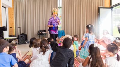 Grethe is wearing massive yellow sunglass and a crown as she tells a story in front of a group of children sitting on the floor