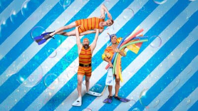 The Dummies stand in front of a blue and white stripey background. One Dummy holds up another over his head, while the third has his arms full of pool noodles and flippers