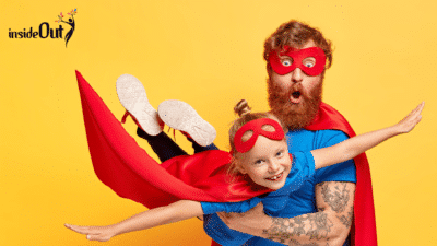 A man dressed as a masked superhero, holds up a young girl, also dressed as a caped superhero, in a flying pose
