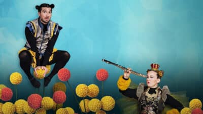 Two performers dressed as bees hang out amongst the flowers. One crouches atop a bloom, while the other spies at him with a telescope