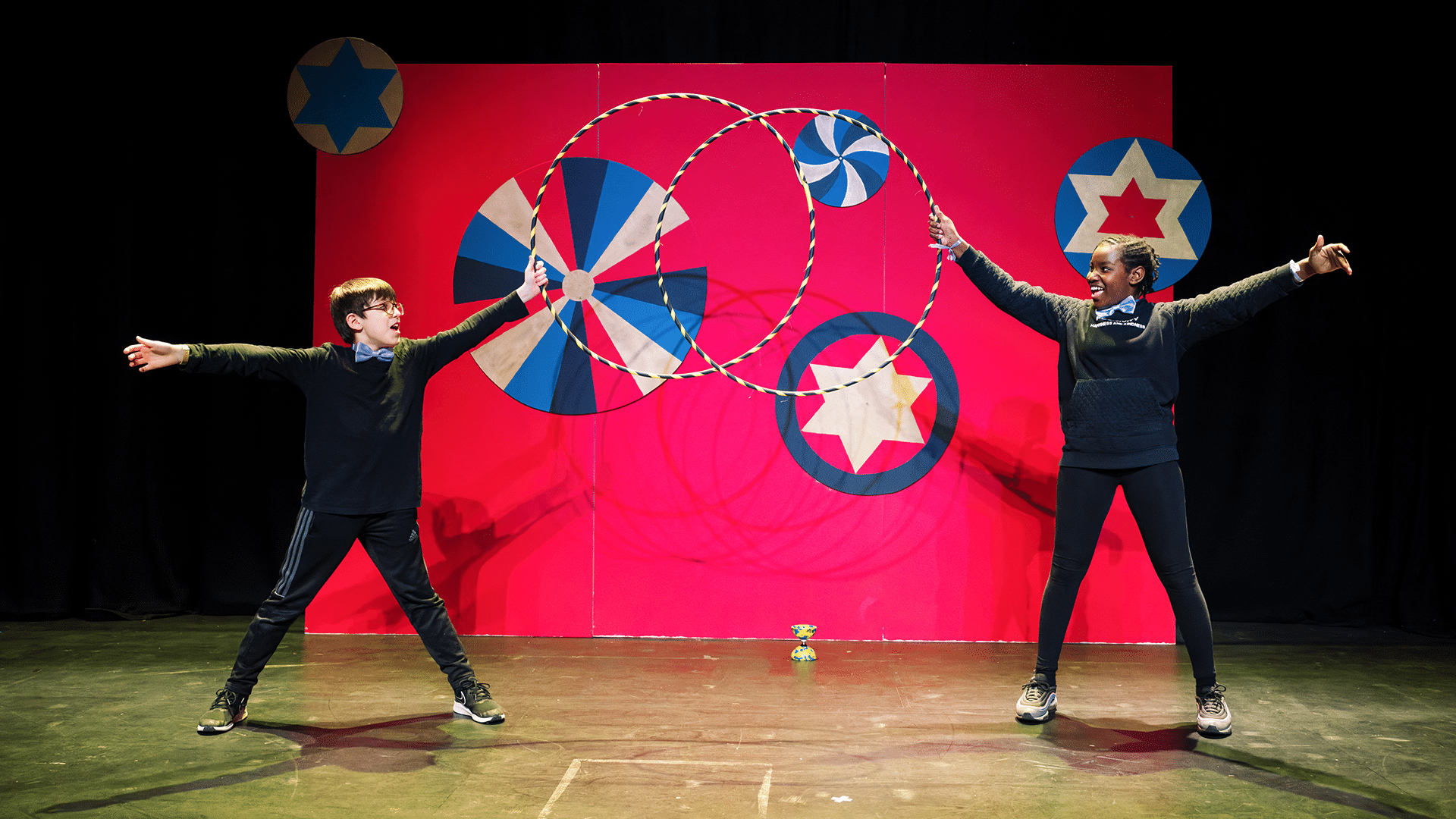 A boy and a girl from Youth Circus hold their final pose with a hula hoop in hand and arms outstretched towards the audience