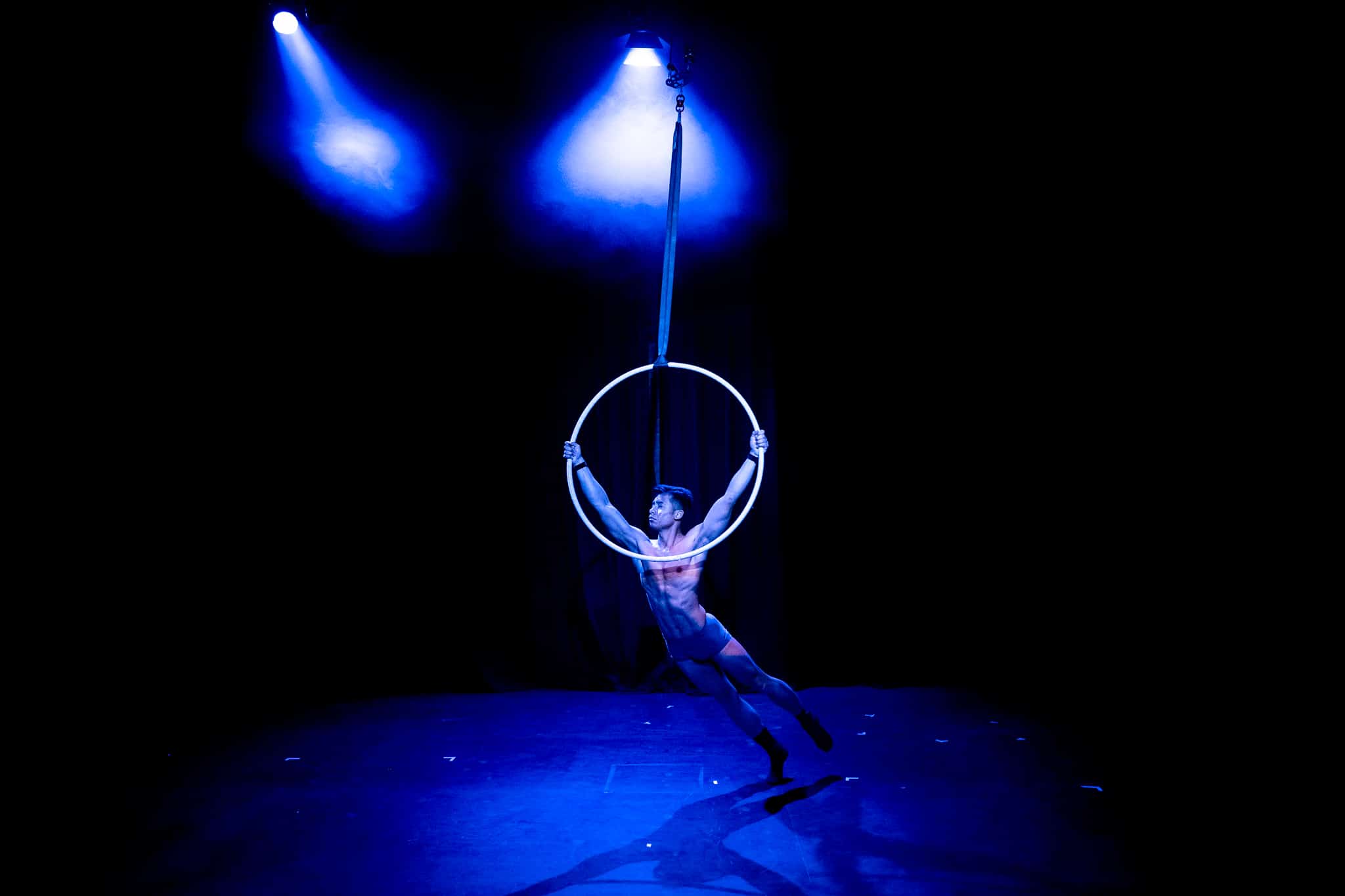 Zaki performing Divinations 1.0 at Jacksons Lane Hangwire Scratch Night (2023). He swings from a hoop with his hands held high and strong in a V shape, his feet barely touching the ground.
