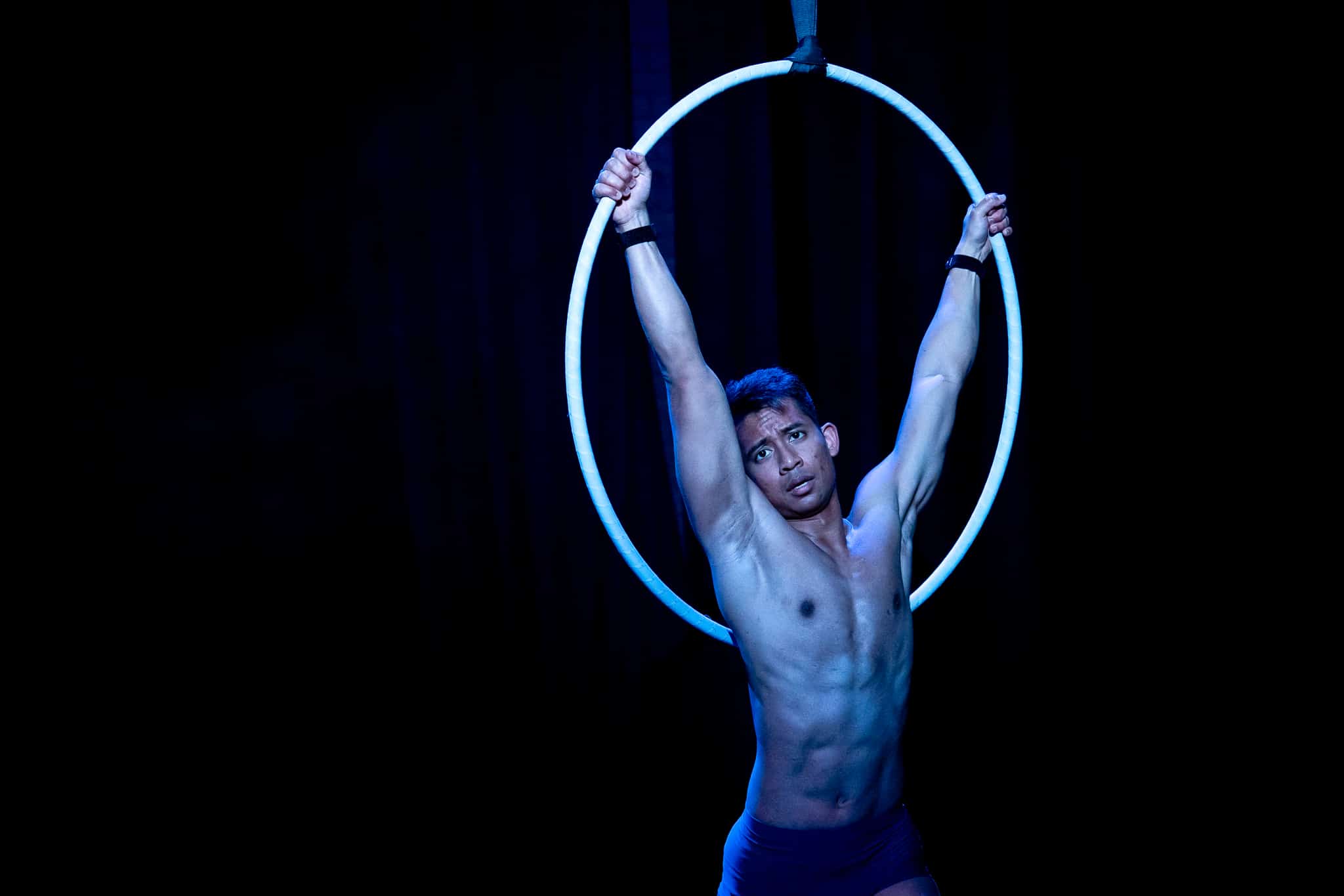 Zaki performing Divinations 1.0 at Jacksons Lane Hangwire Scratch Night (2023). He hangs from a hoop with his hands held high and strong in a V shape, lit with a dim blue light. 
