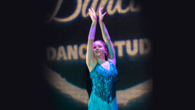 A dancer raises her arms above her head. She wears a turquoise sparkly costume